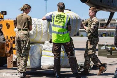 Photo of a person in a UK aid hi-vis vest accompanied by a man and a woman in camouflage clothing loading bundles of humanitarian aid into a plane 