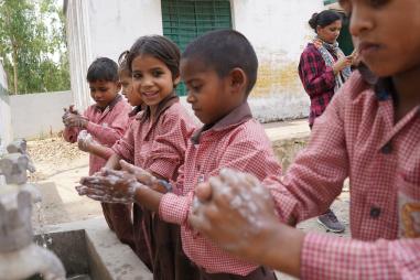 photo of a young girl smiling while she is washing her hands together with her classmates at an outdoor sink in India