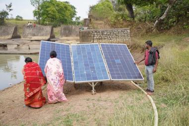 A solar water pump at the edge of a pond in India. Two women dressed in sarees are attending to  the solar panels on the left. There is a man to the right. A group of children watch from a concrete structure in the distance