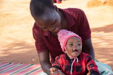 Photo showing a baby sat on their mother's lap on a rug outside in Tanzania. The baby is smiling and the mother is looking lovingly at her child.