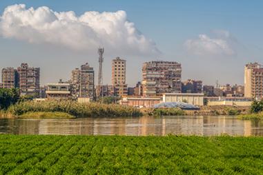 A view of the Nile River flowing between green farmland and modern tall buildings on the other side with a cloudy blue sky, photo from Dahab Island (the golden island) in the middle of the river