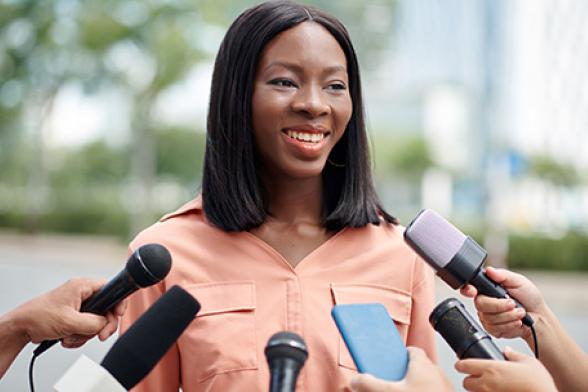 Photo of an African woman speaking at a press conference surrounded by microphones