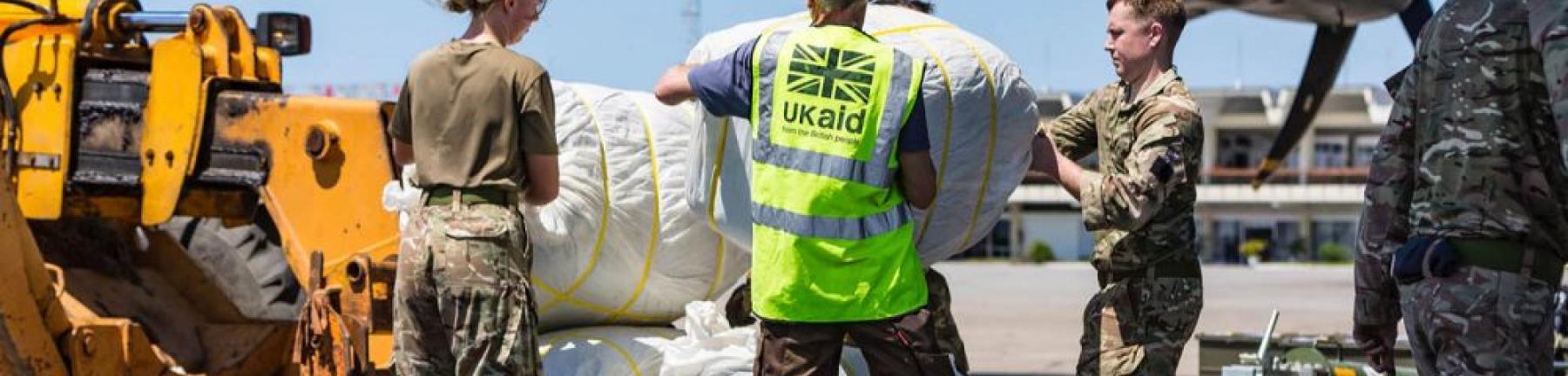 Photo of a person in a UK aid hi-vis vest accompanied by a man and a woman in camouflage clothing loading bundles of humanitarian aid into a plane 
