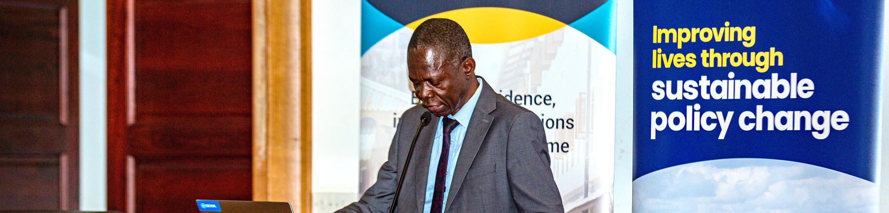 Photo showing Tanzania Country Director, Charles, Sokile, standing at a podium presenting. Behind him are banners, one saying 'deep' and one for Oxford Policy Management with the words 'Improving lives through sustainable policy change' 