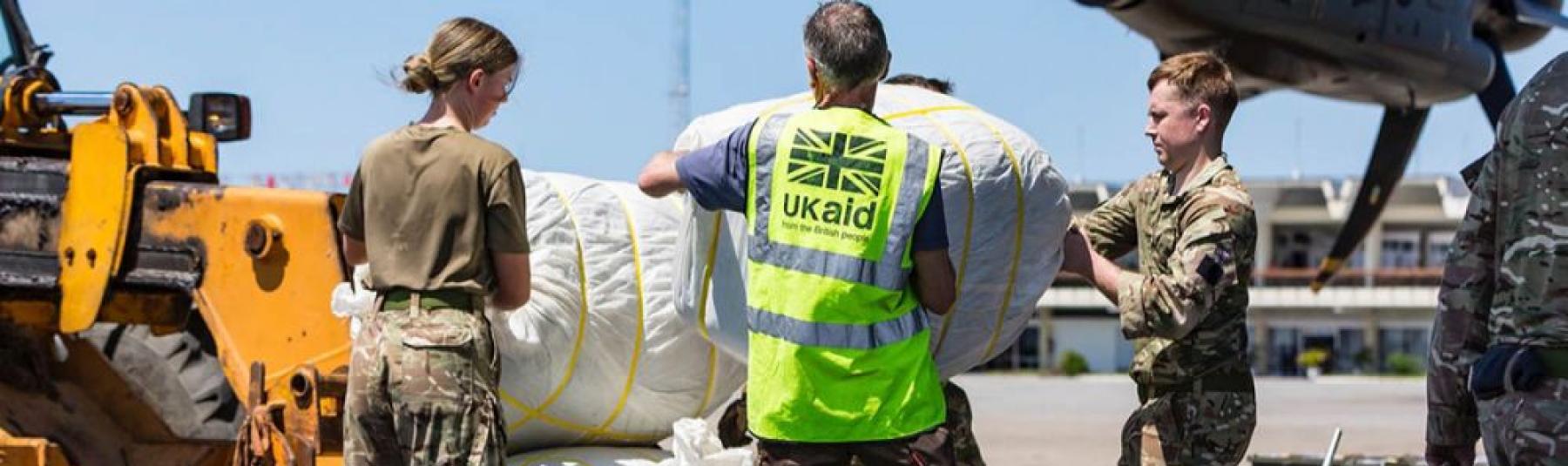 Photo of a person in a UK aid hi-vis vest accompanied by a man and a woman in camouflage clothing loading bundles of humanitarian aid into a plane 