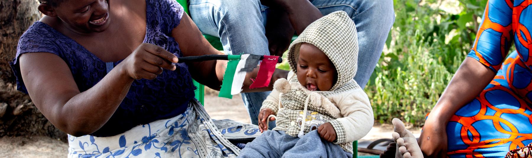 Photo of a mother and baby sat outdoors on a rug in Tanzania. The mother is waving a flag in front of the baby. 