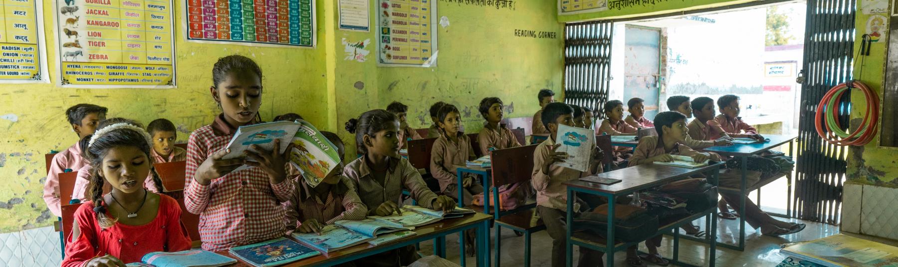 Photo of a classroom in India featuring a mix of children around nine or ten years old.. One girl is standing and reading