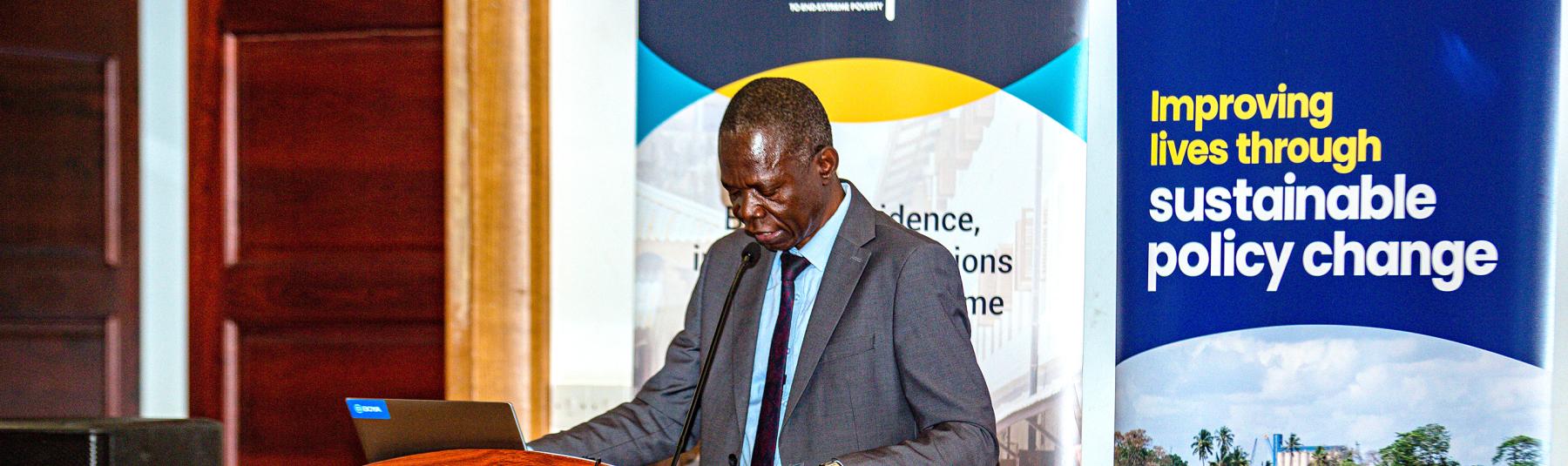 Photo showing Tanzania Country Director, Charles, Sokile, standing at a podium presenting. Behind him are banners, one saying 'deep' and one for Oxford Policy Management with the words 'Improving lives through sustainable policy change' 