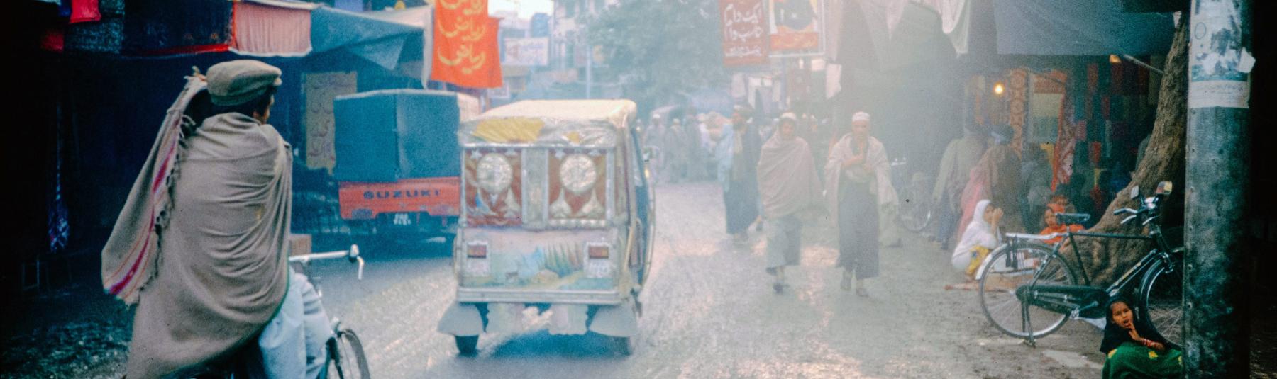 Photo showing a street in Pakistan with a man on a bike, several people walking and a rachshaw